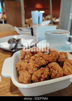 Pollo fritto croccante di popcorn in una ciotola bianca, servito su un tavolo da caffè con sfondo sfocato. Foto Stock