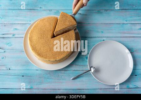 Vista dall'alto di una torta al miele a strati con una fetta sollevata dal server, servita su piatti bianchi su uno sfondo di legno blu. Foto Stock