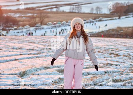 Questa immagine stock mostra una giovane donna vestita con abbigliamento invernale che si gode una bella giornata in un campo innevato. La donna indossa un cappello di pelliccia, A. Foto Stock
