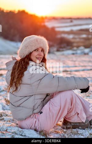 Una giovane donna è seduta sulla neve, vestita con un caldo abbigliamento invernale che include un cappello di pelliccia e un'accogliente giacca a piumino. Si sta godendo un inverno pittoresco Foto Stock