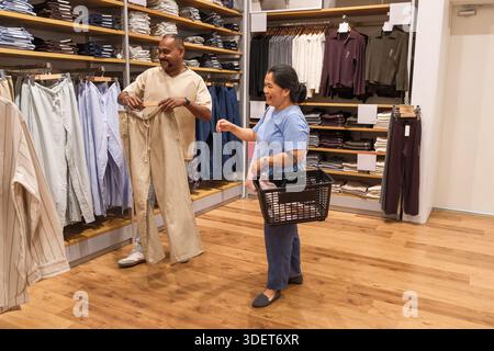 Due persone sono all'interno di un negozio di abbigliamento che guarda i pantaloni e tiene in mano un cestino. Sembrano di buon umore mentre fanno shopping insieme. Foto Stock