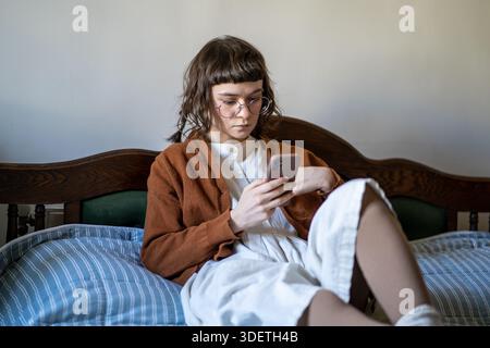 Ragazza hipster pensiva con occhiali in stile vintage che legge le informazioni sullo schermo dello smartphone. Foto Stock