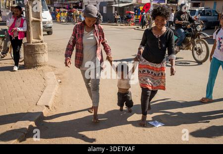 Donne che camminano con un bambino in una strada di Antsirabe, una città degli altopiani centrali del Madagascar. La vita quotidiana nelle aree urbane può essere difficile per le donne che, nonostante l'uguaglianza legale, devono affrontare disparità economiche e norme tradizionali che limitano le opportunità. Molte donne lavorano nel settore informale, vendendo beni o come artigiane, per sostenere le loro famiglie. Quasi un terzo delle donne in Madagascar ha il loro primo figlio prima dei 19 anni. Antsirabe, Madagascar. Foto Stock