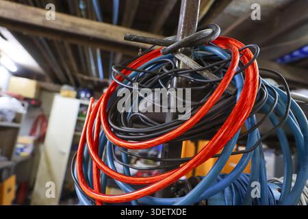 Cavo elettrico avvolto rosso blu e nero appeso su un gancio in metallo arrugginito in un'officina in legno polveroso con sfondo disordinato che mostra uno store disorganizzato Foto Stock