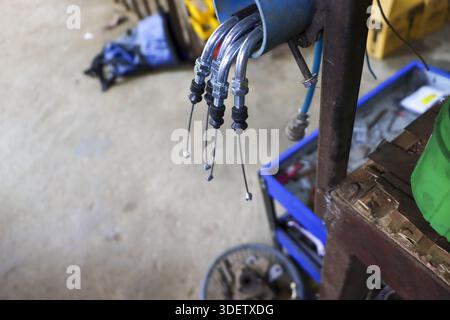 Immagine ravvicinata che guarda verso il basso il lucente cavo metallico per motociclette appeso al banco dei meccanici in un'officina di riparazione affollata, che mostra la manutenzione organizzata Foto Stock