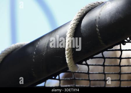 Primo piano, nodo bianco della fune legato intorno alla ringhiera del tubo in acciaio nero pesante che fissa la rete di sicurezza all'esterno sotto la luce del sole che mostra una struttura e una struttura resistenti Foto Stock