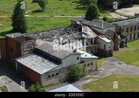 Vista aerea di un edificio industriale abbandonato con un tetto parzialmente crollato circondato da campi verdi e alberi nella campagna della Lettonia. Foto Stock