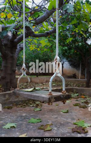 Un vecchio altalena arrugginita è appeso in un giardino vuoto con foglie sparse, una metafora visiva per il tempo, la memoria e la quiete. Foto Stock