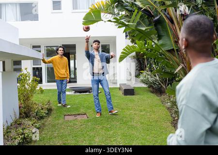 Diversi amici maschili lanciano football americano in un cortile erboso vicino alla casa bianca con bacino d'acqua Foto Stock