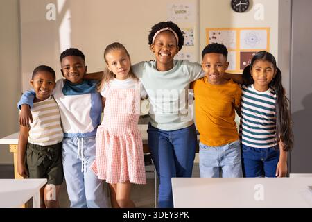 Gruppi diversi di bambini in piedi con le braccia l'uno intorno all'altro, sorridendo in classe vicino alle scrivanie Foto Stock