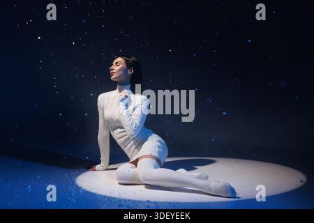 Donna con maglione bianco e calze al ginocchio che si siede contro la neve volante, concetto invernale freddo, Foto Stock