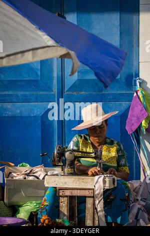 Donna malgascia in un cappello di rafia a tesa larga, seduta e lavorante su una macchina da cucire in stile Singer, Morondava, Madagascar. In Madagascar, le aziende di sartoria spesso operano per strada o appena fuori casa, sfuggendo le linee tra spazio pubblico e privato. Ciò consente alle donne di interagire con i passanti, i potenziali clienti e la comunità generale mentre lavorano. Il lavoro: Le donne in questo ruolo sono abili artigiani, che modificano l'abbigliamento esistente o creano nuovi articoli dai vivaci tessuti lamba popolari nella regione. Foto Stock