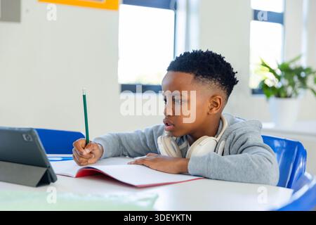 Ragazzo afro-americano che scrive in un notebook rosso in classe, in una felpa con cappuccio grigia, cuffie, utilizzando un tablet Foto Stock