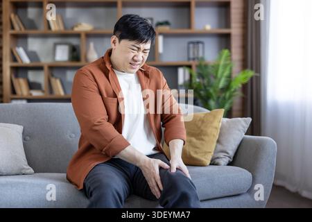 Uomo asiatico che soffre di grave dolore al ginocchio mentre seduto su un divano grigio a casa, toccando la gamba in disagio e sembra angosciato dall'articolazione inj Foto Stock