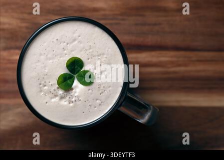 Colpo ad angolo alto di una tazza di birra scura con punta in schiuma cremosa decorata con un trifoglio verde su una superficie in legno. Il giorno di San Patrizio Foto Stock