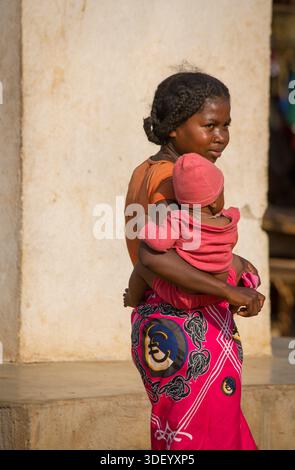 Giovane madre malgascia che porta il suo bambino, Bekopaka, Madagascar. Il villaggio di Bekopaka è un remoto villaggio nella regione malgascia del Madagascar occidentale e funge da punto di accesso principale e base logistica per visitare il famoso Parco nazionale di Tsingy de Bemaraha. La comunità nel villaggio di Bekopaka è costituita principalmente dal gruppo etnico Sakalava, che sono gli abitanti tradizionali di questa regione del Madagascar occidentale. La vita della comunità è profondamente radicata nelle tradizioni locali, nella venerazione degli antenati e in un mix di mezzi di sussistenza tradizionali e turismo fiorente. Foto Stock
