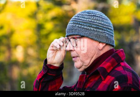 Un uomo anziano con un cappello a maglia e occhiali adatta i suoi occhiali all'aperto, circondato da una vegetazione lussureggiante, catturando un momento sereno di riflessione e natura Foto Stock