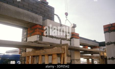 Cantiere con una gru che solleva un blocco di cemento su un muro parzialmente costruito, unendo mattoni rossi e travi prefabbricate Foto Stock