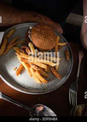 Vista dall'alto di un piatto con un hamburger di semi di sesamo e patatine fritte sparse su un tavolo di legno scuro - ombre foto moody Foto Stock