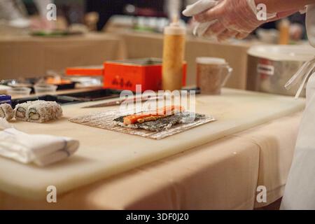 Un sushi chef sta preparando un sushi roll su un tagliere. Il sushi roll è preparato con un pezzo di salmone e alcune verdure. Lo chef sta usando un coltello Foto Stock