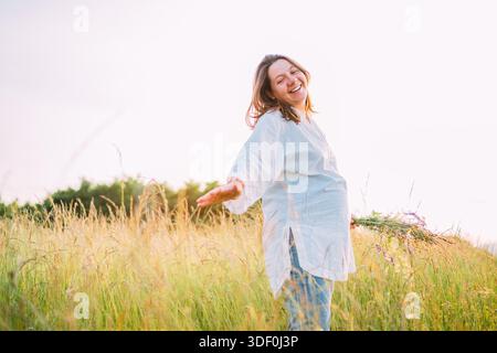 Sorridendo sinceramente, giovane donna incinta vestita con abiti estivi leggeri, che cammina su un prato di erba verde con bouquet di fiori selvatici durante il sole serale Foto Stock
