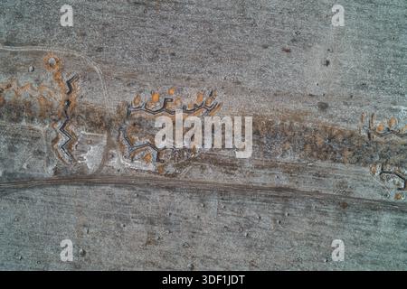 Una linea di trincea militare a zigzag taglia attraverso il suolo ghiacciato parallelo a una strada di campagna, creando una barriera difensiva. Questa vista aerea invernale mette in risalto i lavori tattici in terra e la logistica strategica di una zona di combattimento. Foto Stock