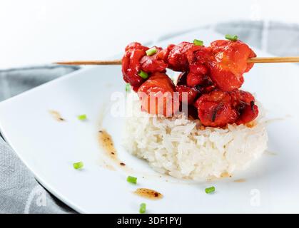 Spiedini di pollo rosso conditi serviti su un piccolo mucchio di riso bianco appiccicoso al vapore su un piatto quadrato bianco. Cucina asiatica o fusion. Foto Stock