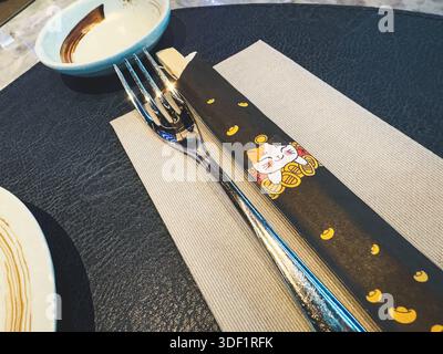 Roma, italia 2 gennaio 2026: Cena a tavola in un ristorante giapponese o asiatico, con forchetta e bacchette con un gatto maneki neko tradizionale Foto Stock