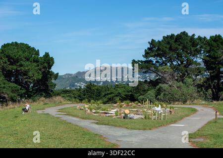 Un parco tranquillo caratterizzato da un percorso tortuoso che conduce attraverso una vegetazione lussureggiante, con una piccola disposizione in pietra nel centro. Sullo sfondo, colline e. Foto Stock