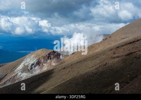 Una vista panoramica di un paesaggio vulcanico caratterizzato da una catena montuosa con vapore che sale dalle ventole vulcaniche. Il cielo è parzialmente nuvoloso, creando un dramma Foto Stock