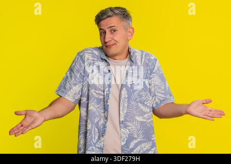 Uomo di mezza età che si scaglia le spalle e alza le mani con un aspetto incerto, esprimendo confusione e mancanza di risposte. Giovane ragazzo che mostra emozioni perplesse e reazioni poco chiare isolato su sfondo giallo Foto Stock