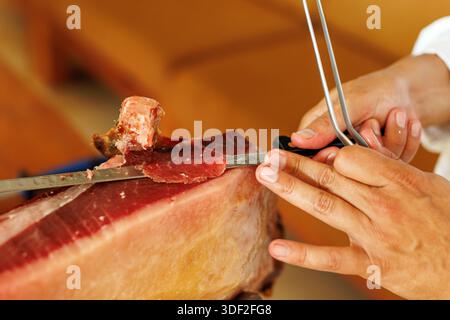 Primo piano di mani che affetta il prosciutto stagionato con un lungo coltello da intaglio, mostrando il processo di taglio preciso. Foto Stock