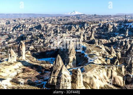 Camini fatati coperti di neve si ergono dal paesaggio invernale vicino a Göreme nella Cappadocia. In lontananza si staglia lo stratovulcano Erciyes Dağı (3.917 m/12.851 ft), la montagna più alta dell'Anatolia centrale. Con le sue formazioni erose di roccia tuffacea, la Cappadocia fa parte del sito del patrimonio mondiale UNESCO "Parco nazionale di Göreme e i siti rupestri della Cappadocia" dal 1985. Turchia. Foto Stock