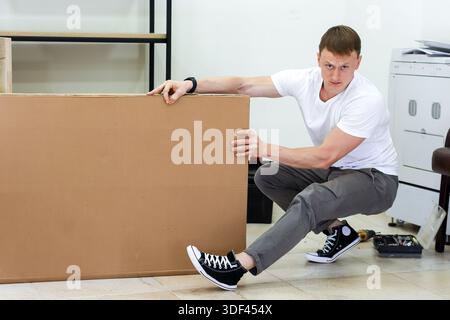 Uomo che si impegna a sollevare e spostare un pannello di cartone grande. Giovane atletico che solleva e porta con sé un foglio di cartone marrone oversize in stile moderno Foto Stock
