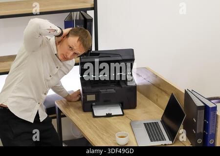 Il dipendente in ufficio sopraffatto gratta la testa nella confusione mentre si appoggia alla stampante rotta nell'ambiente di lavoro. Foto Stock