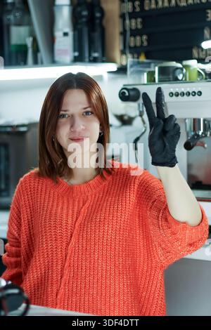 Una lavoratrice in guanto posa con due dita in alto in un moderno ambiente da caffetteria Foto Stock
