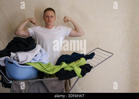 Il giovane uomo muscoloso in t-shirt bianca si flette bicipite accanto a un rack carico di biancheria in vari colori e un cesto blu su sfondo beige Foto Stock