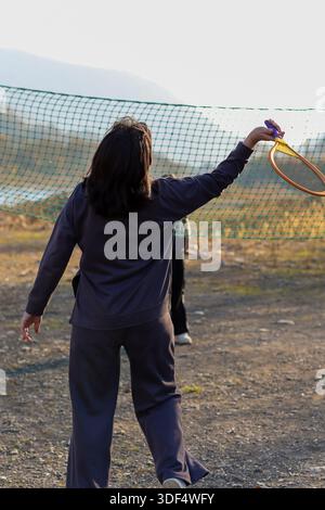 Donna dinamica in un outfit grigio sportivo che si distingue con una racchetta da tennis sopra la rete sul campo aperto e sullo splendido paesaggio montano. Foto Stock