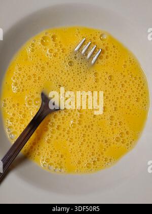 Uova crude sbattute in un recipiente bianco dalla consistenza spumosa e gialla con una forchetta di metallo per sbattere la colazione fresca fatta in casa o preparare la cottura, Foto Stock