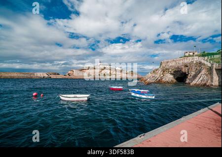 Il porto di Tapia de Casariego sul Mar Cantabrico presenta piccole barche da pesca ormeggiate in acque blu profonde, con il faro della città che si affaccia sulla roc Foto Stock