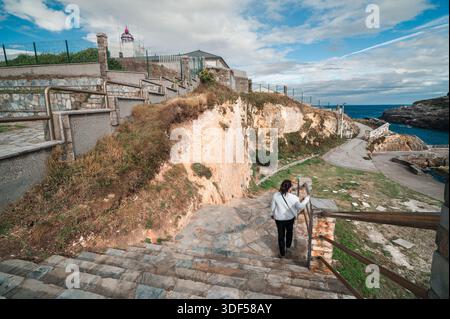 Donna che scende le scale di pietra lungo una costa frastagliata, che conduce a un porto panoramico con un faro storico visibile sulla cima della scogliera sotto una nuvolosa S. Foto Stock