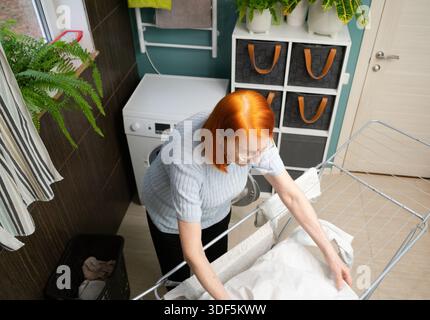 Una prospettiva dall'alto verso il basso mostra una donna anziana che prepara con cura abiti bagnati e appena lavati su un rack di asciugatura in metallo pieghevole in casa sua. Foto Stock