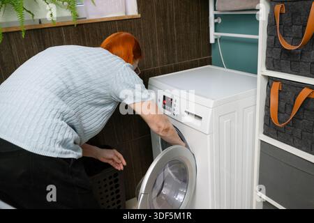 Una prospettiva posteriore cattura una donna anziana con un mal di schiena, scaricando un bucato pulito bagnato da una lavatrice a carico frontale. Foto Stock