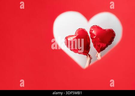 Due mani reggono palloncini rossi a forma di cuore attraverso una fessura a forma di cuore su sfondo rosso, amore reciproco, San Valentino Foto Stock