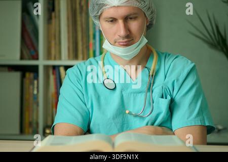 Un medico o un operatore medico in uniforme con maschera di protezione legge un libro alla sua scrivania. Stetoscopio sul collo. Concetto di educazione medica, sviluppo professionale Foto Stock