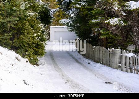 Vialetto coperto di neve fiancheggiato da una recinzione in legno usurata che conduce a un garage in una soleggiata giornata invernale Foto Stock