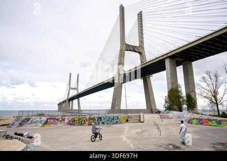 Lisbona Portogallo 18 dicembre 2025. Le persone viaggiano in bicicletta e skateboard in uno skatepark in cemento sotto il ponte Vasco da Gama lungo il fiume Tago. Foto Stock