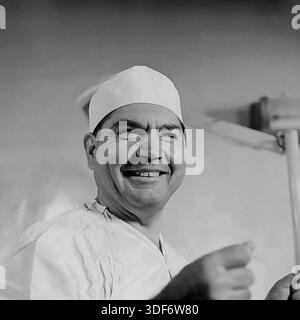 Slaviansk, RSS Ucraina, 1970S.. Un emozionante ritratto storico da vicino di un chirurgo maschile sorridente con un berretto di cotone bianco. Il medico esperto sta ridendo, guardando da parte con un'espressione allegra che contrasta con la solita tensione del suo lavoro. Le sue mani sfocate suggeriscono un gesto vivace. Lo sfondo mostra una lampada chirurgica. Questa autentica fotografia d'archivio monocromatica cattura il lato umano, il sollievo e l'ottimismo degli operatori sanitari sovietici nella pacifica regione del Donbass. Foto Stock