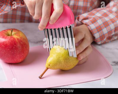 Un bambino affetta una pera con un coltello in acciaio inossidabile ondulato su un tagliere rosa, primo piano Foto Stock