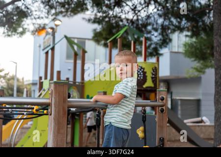 Ragazzo biondo in camicia a righe in piedi sulla piattaforma del parco giochi tenendo in mano le ringhiere e guardando la macchina fotografica nel parco del resort con strutture colorate. Concetto di Foto Stock